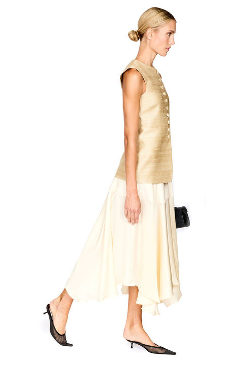A woman in profile wearing a sleeveless champagne-gold vest and a cream-colored layered skirt, paired with black pointed-toe mule heels, against a solid white background.