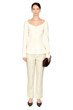 A model stands against a plain white background wearing a matching cream-colored two-piece suit consisting of a long-sleeve top with a wide scoop neckline and wide-leg trousers. She is holding a small dark brown leather clutch and wearing dark pointed-toe flats.