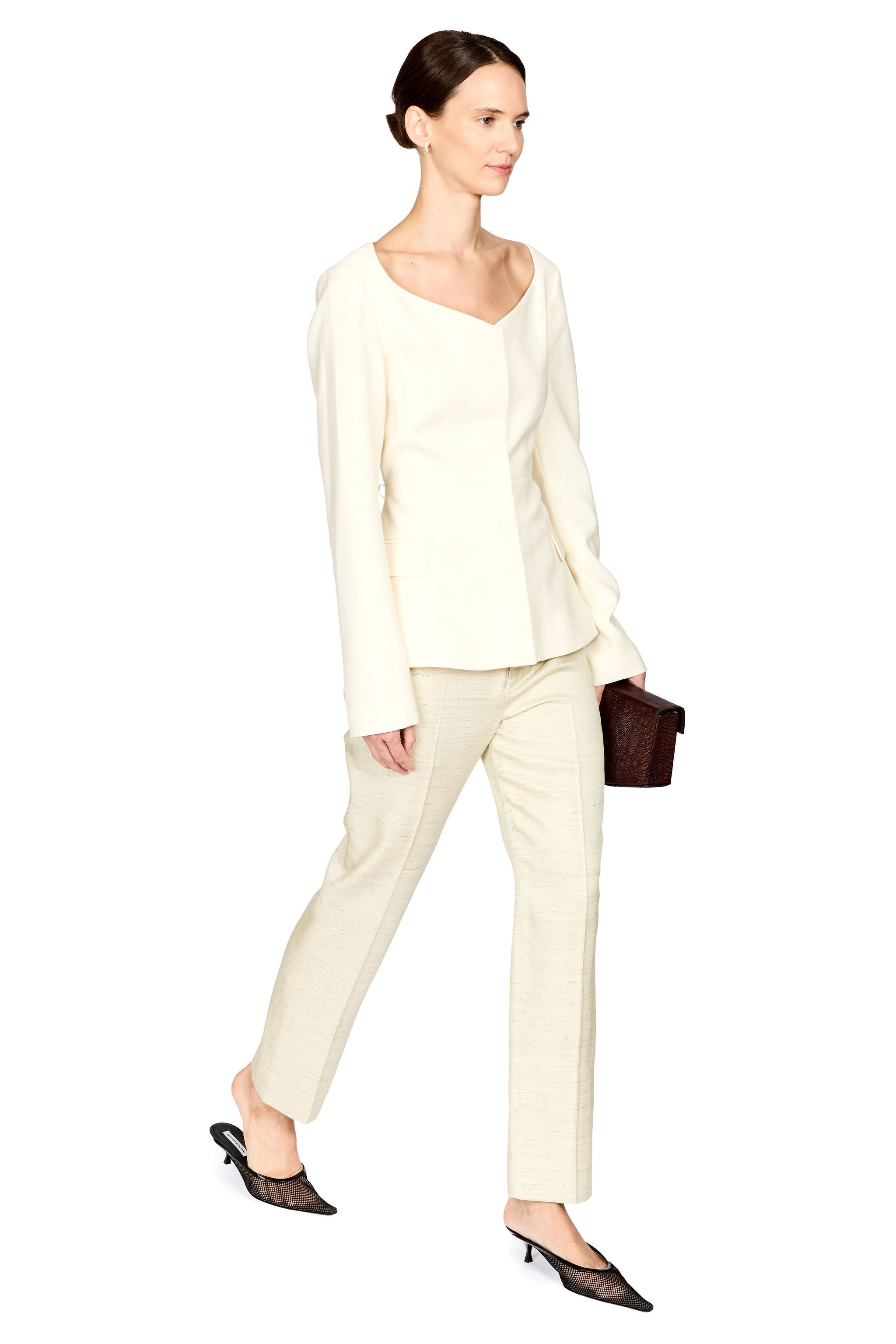 A woman modeling a cream-colored two-piece set consisting of a long-sleeve V-neck top and matching straight-leg trousers, styled with black pointed-toe mule heels and a small dark brown handbag against a solid white background.