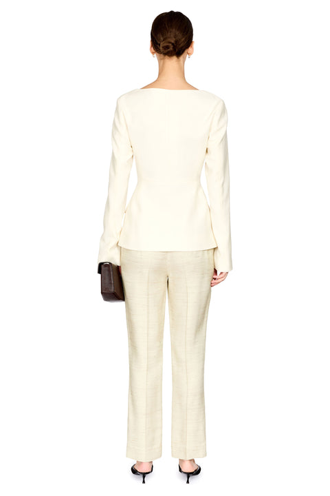 A woman shown from the back wearing a cream-colored long-sleeve top and matching cream trousers, holding a small dark brown handbag against a plain white background.