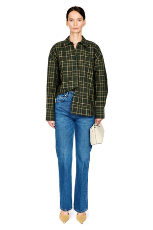 A model stands against a white background wearing a dark green and black plaid button-up shirt, blue jeans, beige pointed shoes, and holding a small cream-colored handbag.