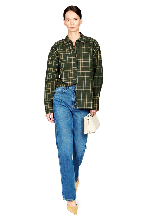 A model wearing a green and black plaid button-up shirt, blue jeans, beige heels, and holding a small cream handbag against a solid white background.