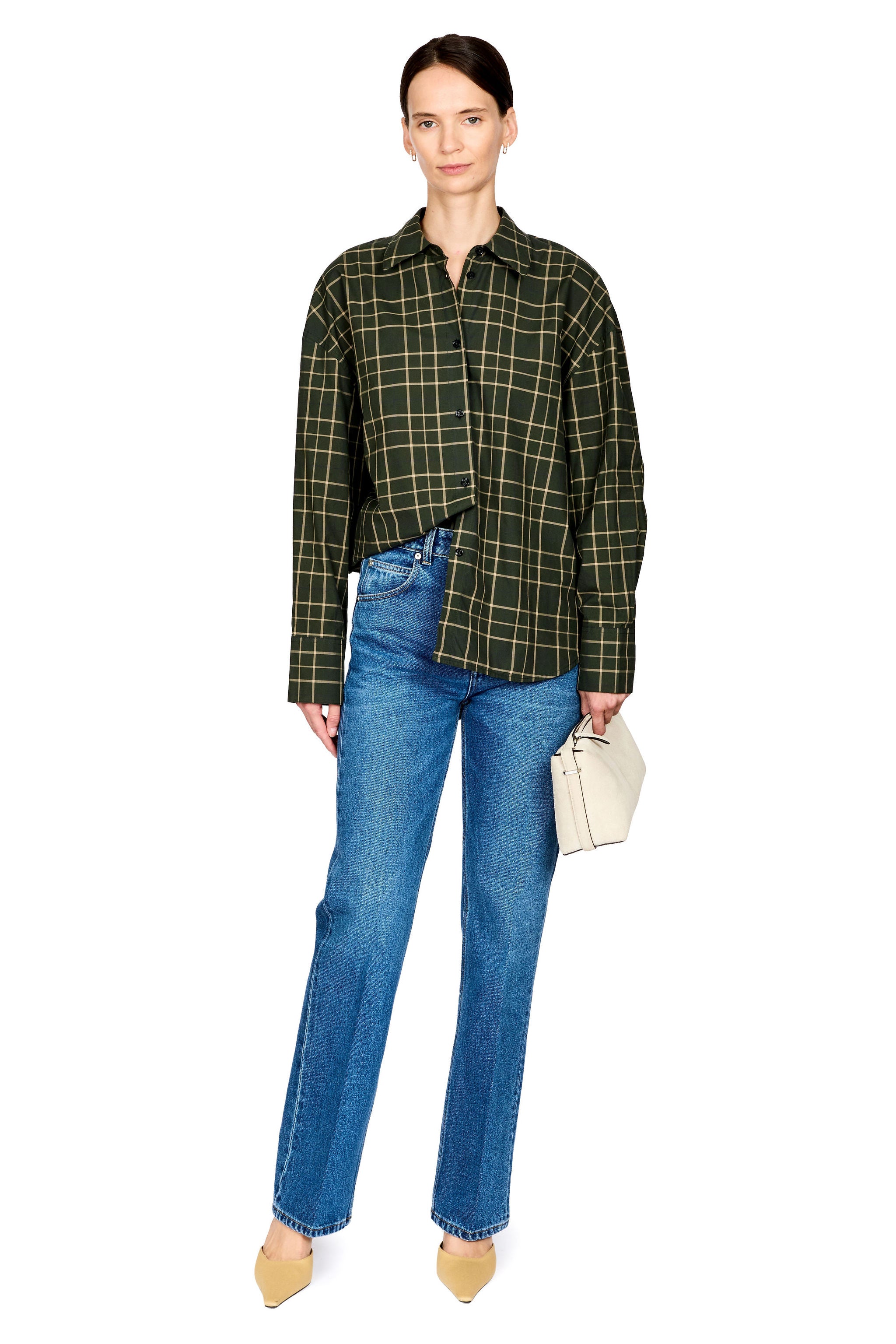 A model stands against a white background wearing a dark green and black plaid button-up shirt, blue jeans, beige heels, and holding a small cream-colored handbag.
