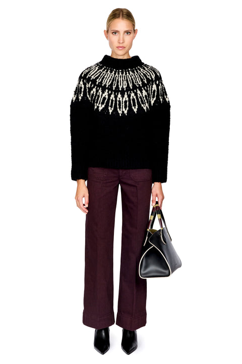 A model stands against a white background wearing a black knit sweater with a white geometric pattern across the shoulders and upper chest, paired with dark burgundy wide-leg trousers and black pointed shoes, while holding a black handbag.
