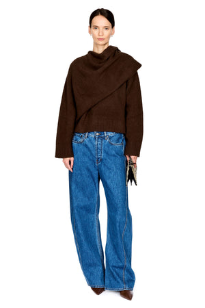 A model wearing a dark brown asymmetrical cowl-neck knit sweater and wide-leg blue jeans, holding a small black clutch against a plain white background.