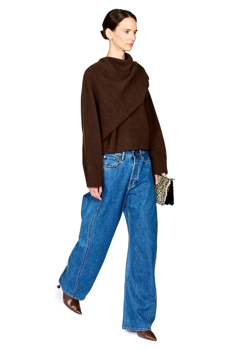 A model wearing a dark brown asymmetrical cowl-neck knit sweater, wide-leg blue jeans, and brown pointed-toe boots, holding a small patterned clutch against a plain white background.