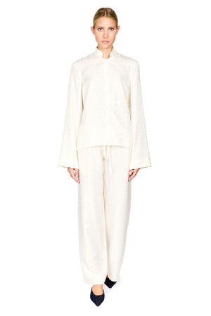 A woman modeling a matching cream-colored two-piece outfit consisting of a long-sleeve cropped jacket with a mandarin collar and wide-leg trousers, set against a plain white background.