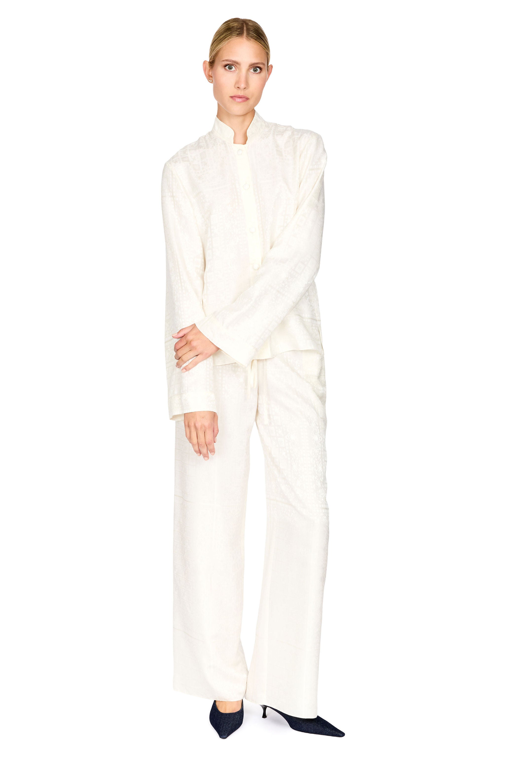 A model wearing a matching off-white textured two-piece set consisting of a long-sleeve mock-neck top and wide-leg trousers, styled with dark pointed-toe heels against a plain white background.