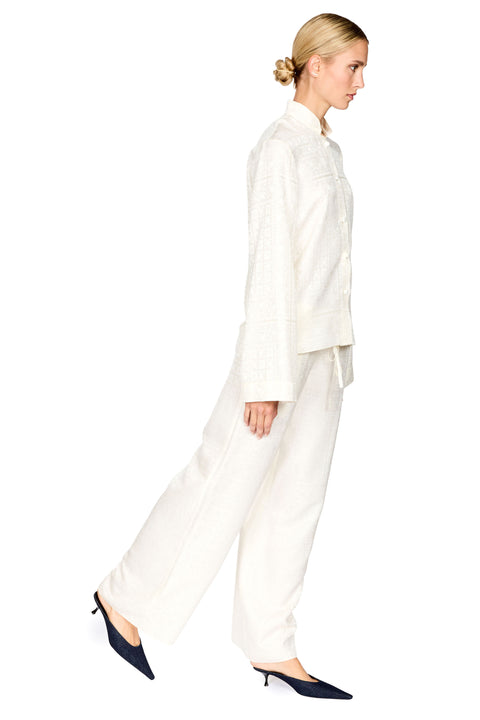 A woman modeling a matching white textured two-piece set consisting of a long-sleeve button-front jacket and wide-leg trousers, paired with dark navy pointed-toe heels against a plain white background.
