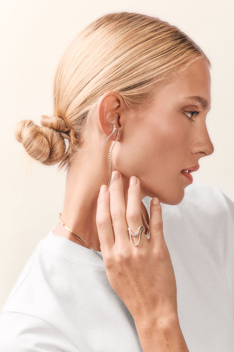 A blonde woman in profile wearing delicate gold jewelry, including a long curved earring, a thin chain necklace, and stacked rings on her hand, against a soft cream background.