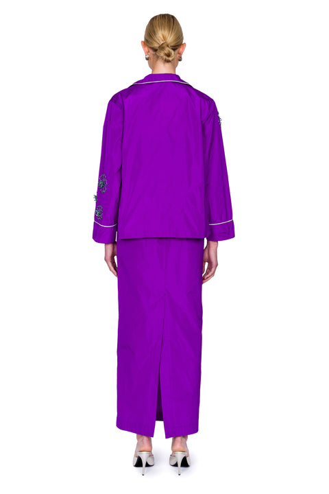 Back-facing woman wearing a purple button down pajama shirt with floral embellishments and a matching maxi skirt.