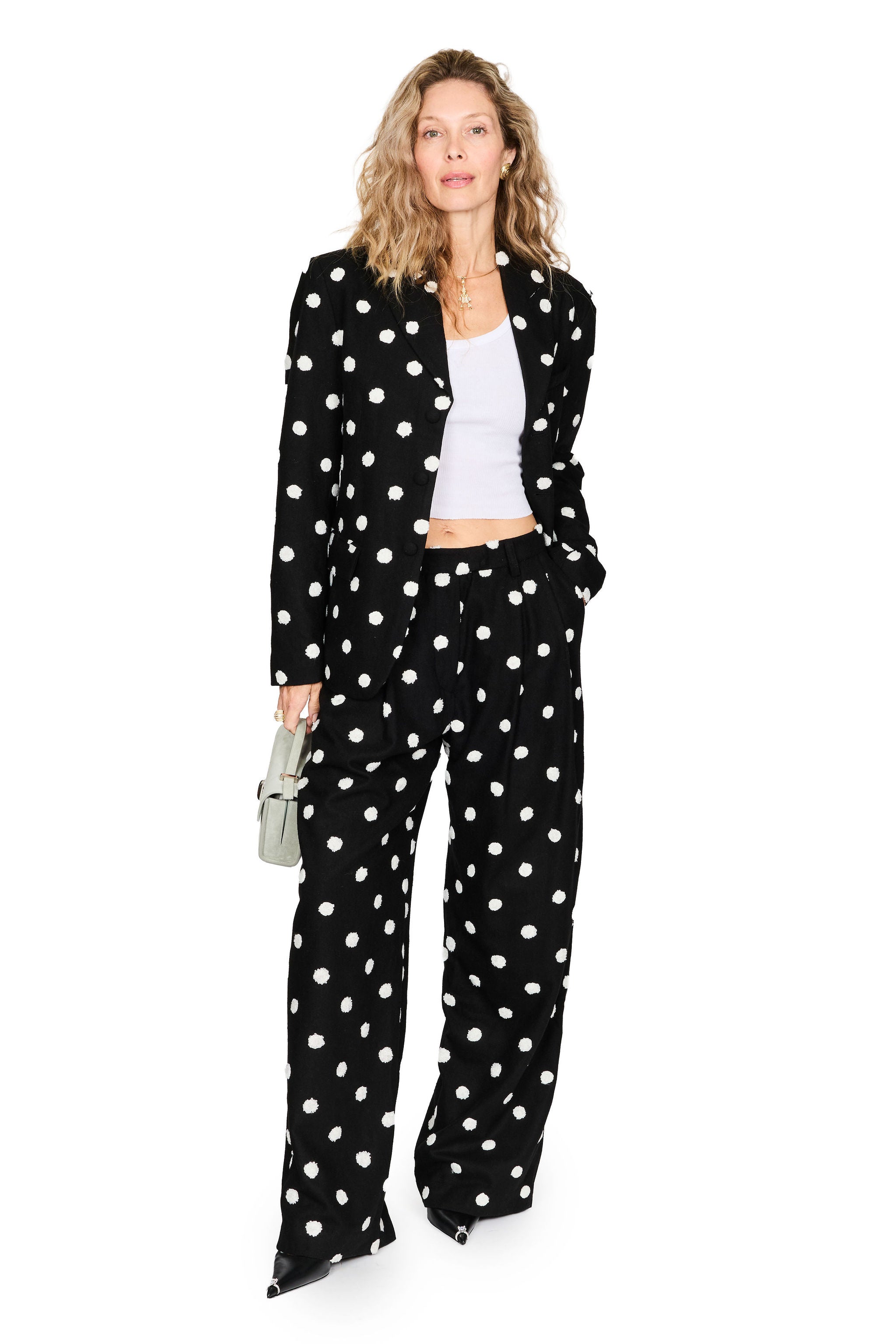 A woman modeling a black velvet two-piece pantsuit with white polka dots, consisting of a blazer and wide-leg trousers, styled with a white crop top and holding a small gray handbag against a white background.