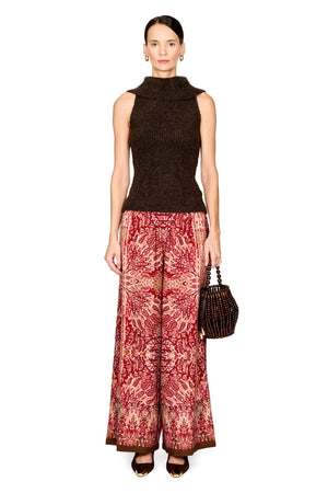 A model stands against a white studio background wearing a sleeveless dark brown knit mock-neck top, wide-leg red and beige patterned trousers, and holding a small dark woven handbag.