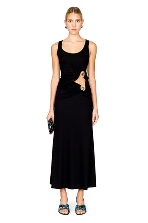 A model stands against a white background wearing a sleeveless black top and maxi skirt with a draped waist detail and a gold-toned ring accent. She is holding a small black patterned clutch and wearing blue patterned flat sandals.