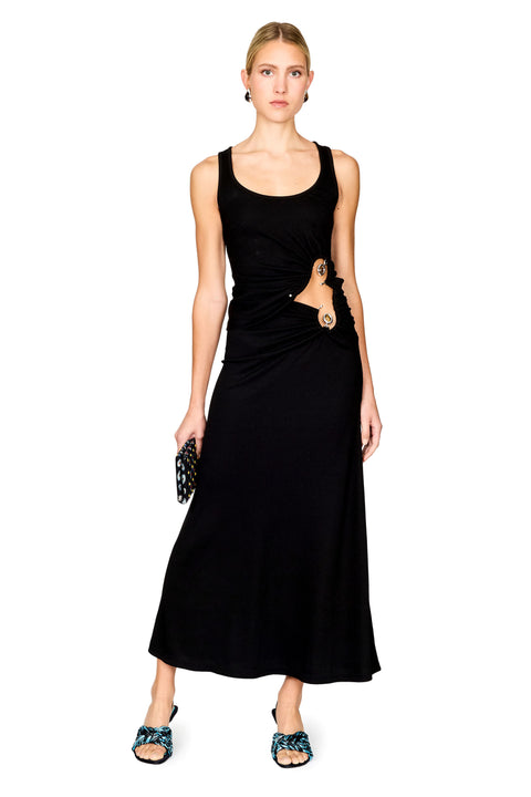 A model stands against a white background wearing a sleeveless black top and maxi skirt with a draped waist detail and a gold-toned ring accent. She is holding a small black patterned clutch and wearing blue patterned flat sandals.