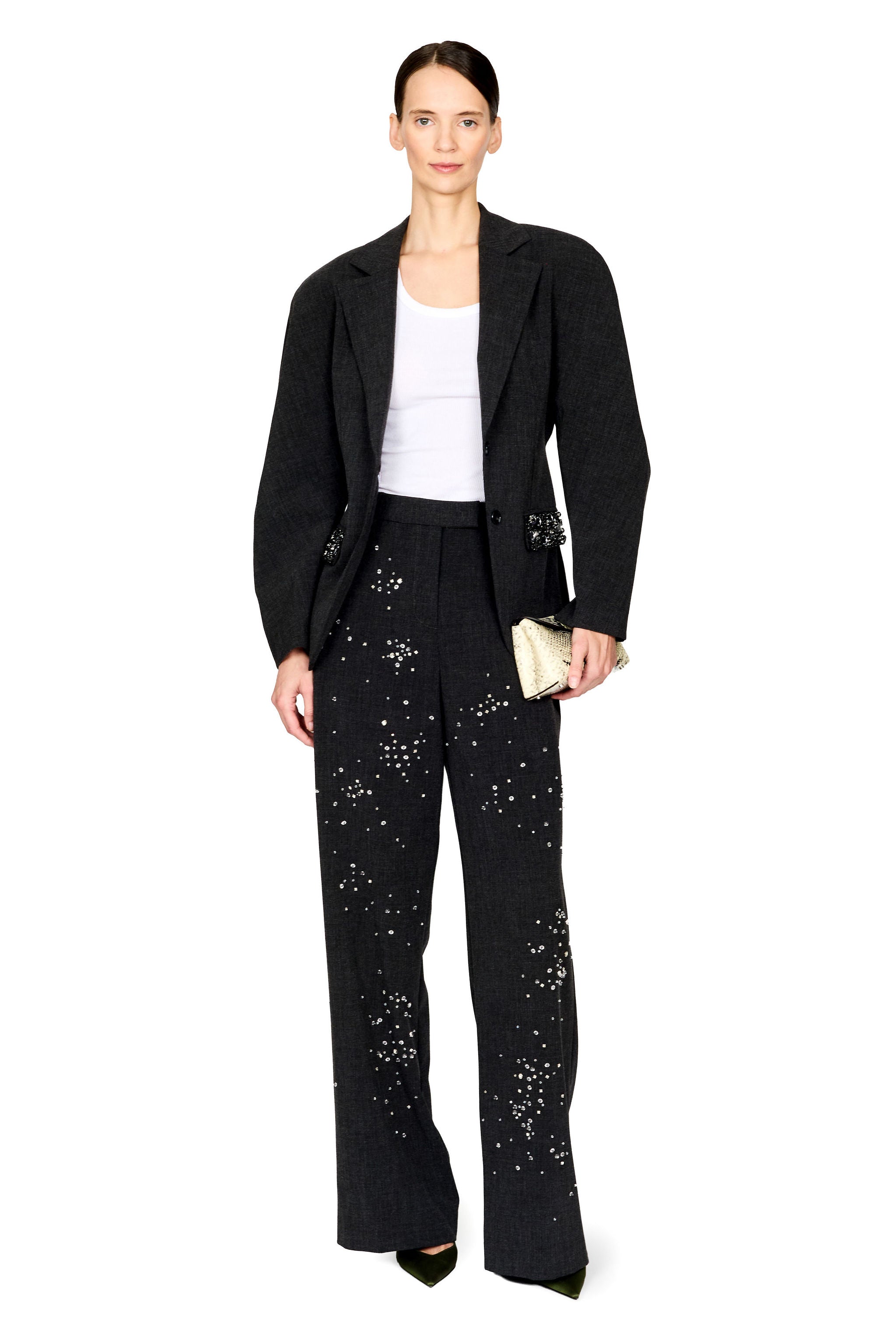 A model stands against a white studio background wearing a black textured blazer and matching wide-leg trousers with scattered silver embellishments, paired with a white top and a small metallic clutch.
