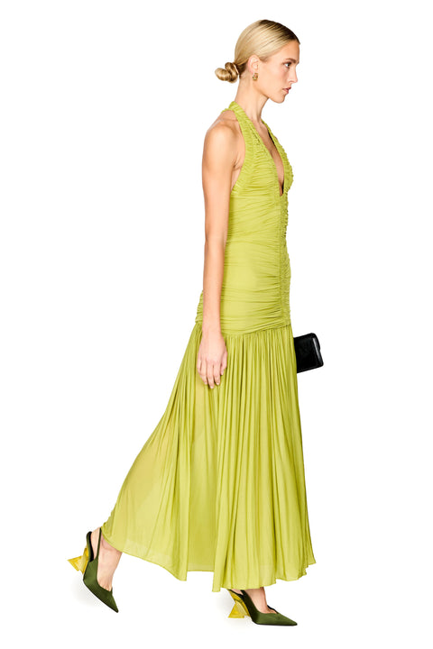 A woman modeling a sleeveless halter-neck maxi dress in a bright yellow-green, shown from the side against a plain white background. She is holding a small black clutch and wearing olive green pointed-toe heels.