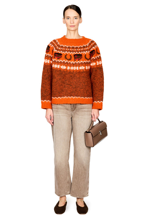 A model stands against a white studio background wearing an orange and brown patterned knit sweater, beige wide-leg jeans, dark brown flats, and holding a brown handbag.
