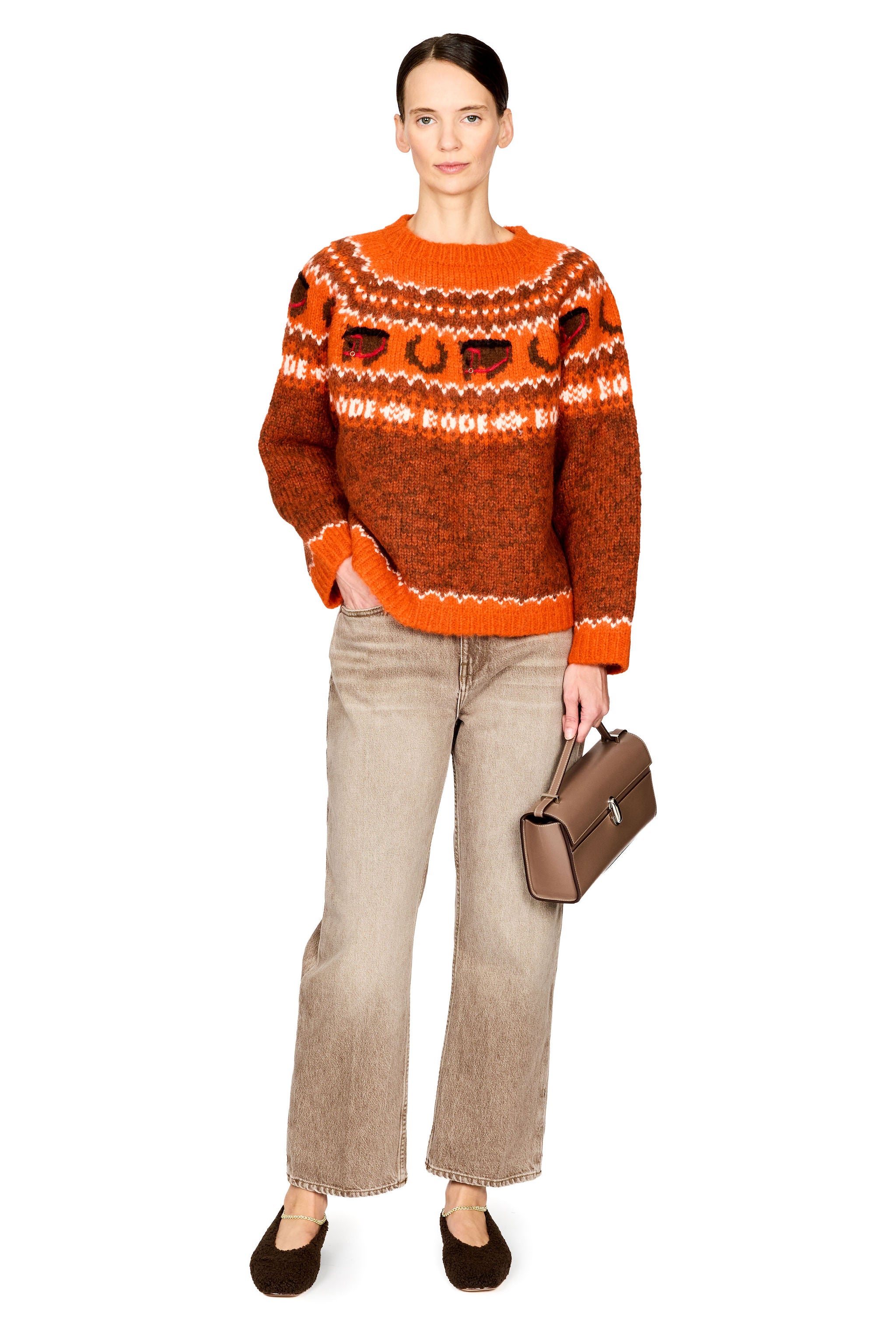 A woman stands against a plain white background wearing a bright orange and white patterned knit sweater, beige wide-leg jeans, dark brown flats, and holding a brown handbag.