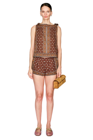 A model stands against a white background wearing a sleeveless brown patterned top and matching shorts, holding a small tan clutch bag.