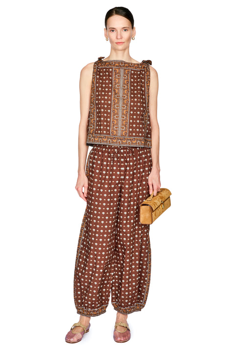 A model stands against a white studio background wearing a brown sleeveless top and matching wide-leg pants with a white polka dot pattern, holding a small tan leather clutch.