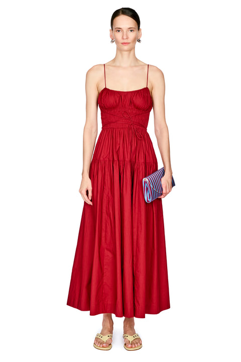 A woman modeling a long red spaghetti-strap dress with a ruched bodice and a flared skirt, holding a blue and red striped clutch against a plain white background.