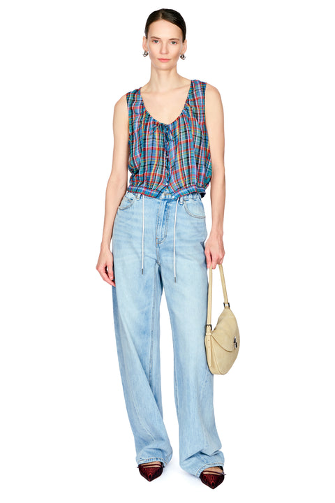 A model stands against a white studio background wearing a colorful plaid sleeveless top, light-wash wide-leg denim drawstring pants, dark pointed shoes, and holding a beige crescent handbag.