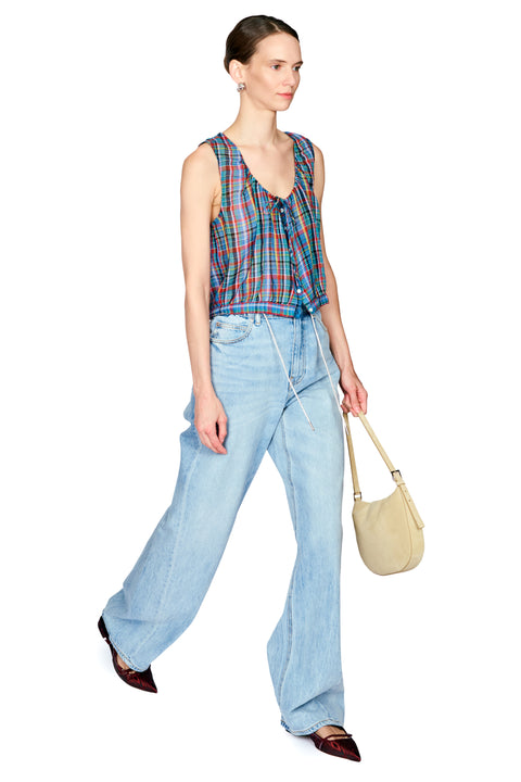 A model is shown from the head to toe wearing a colorful plaid sleeveless top, light-wash wide-leg jeans, and dark brown pointed flats, while holding a beige crescent shoulder bag against a plain white background.