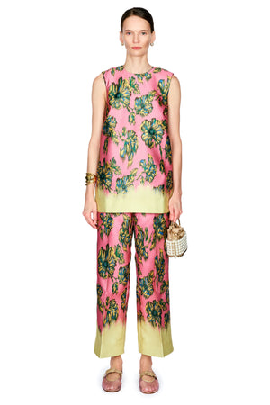 A model stands against a white background wearing a sleeveless pink top with a green and blue floral print and a yellow-green hem, paired with matching wide-leg pants, a gold cuff , and a small woven handbag.