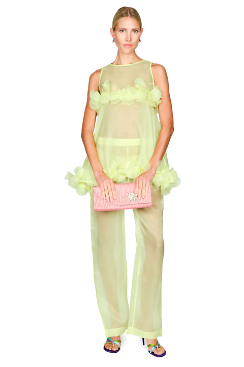 A model wearing a sheer pale yellow-green ruffled sleeveless top and matching wide-leg pants, holding a pink quilted clutch bag, against a plain white background.