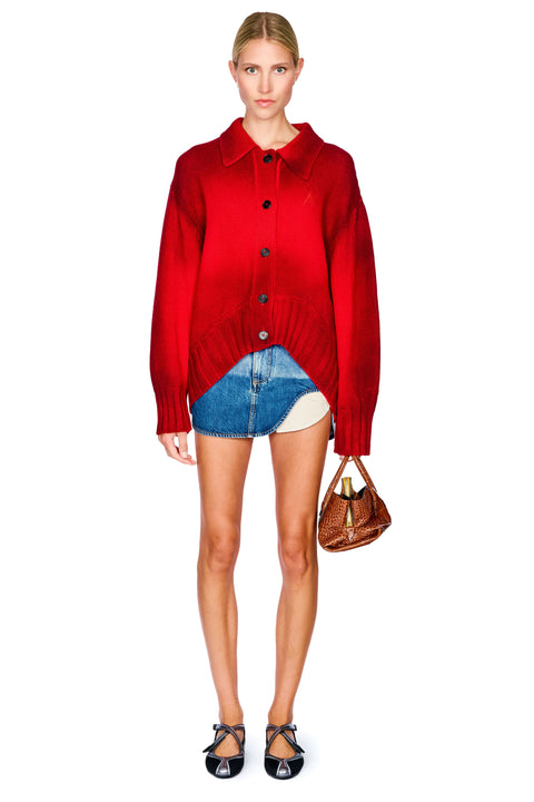 A model stands against a white background wearing a bright red button-up knit cardigan, a denim mini skirt with an asymmetrical hem, black strappy heels, and holding a brown handbag.