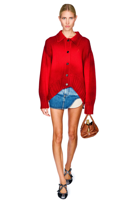 A model stands against a white background wearing a bright red button-up knit cardigan, a denim mini skirt with an asymmetrical hem, black strappy heels, and holding a brown handbag.