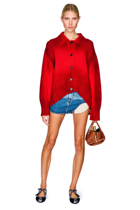 A model stands against a white background wearing a bright red button-front cardigan over a denim mini skirt, holding a brown handbag.