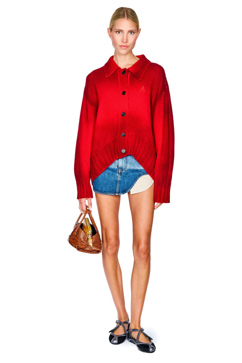 A model stands against a white background wearing a bright red button-up knit cardigan, a denim mini skirt with an asymmetrical hem, black strappy heels, and holding a brown handbag.
