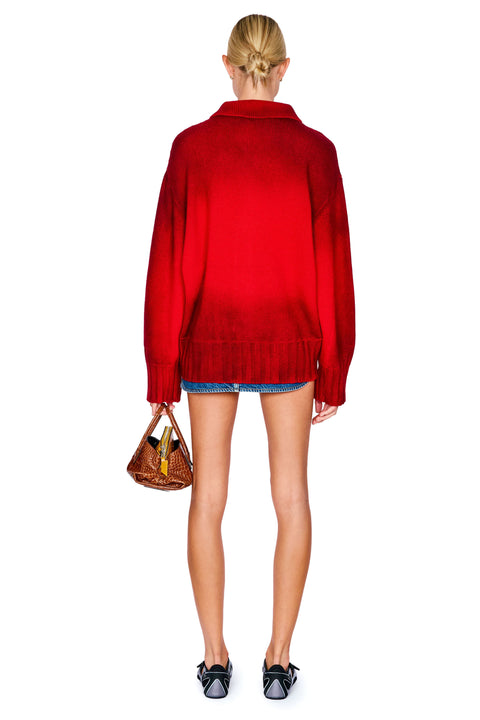 A model shown from the back wearing a vibrant red knit cardigan, a denim mini skirt, black shoes, and holding a brown woven handbag against a plain white background.