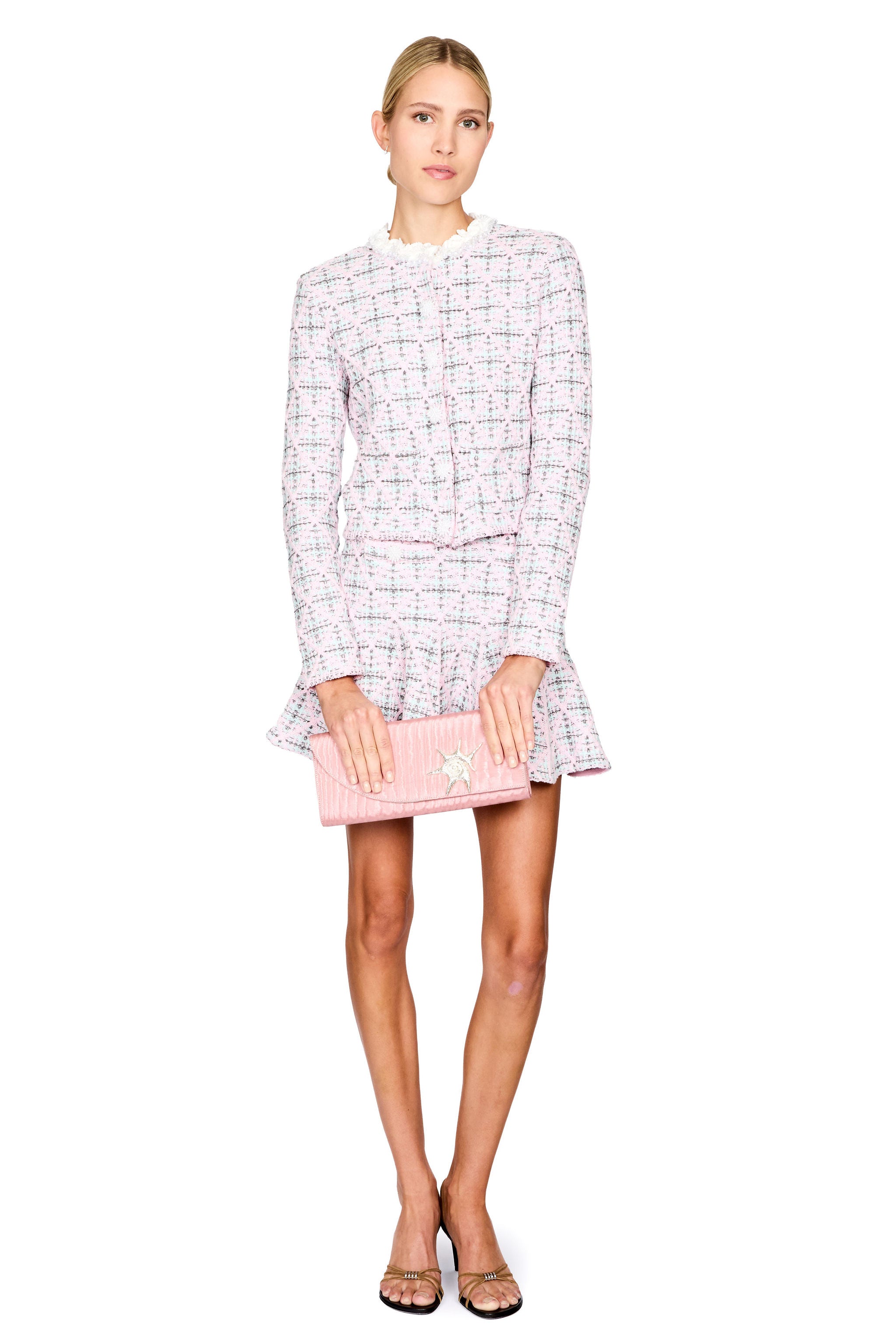 Model stands against a white background wearing a light pink and white tweed jacket and skirt set, holding a pink clutch bag